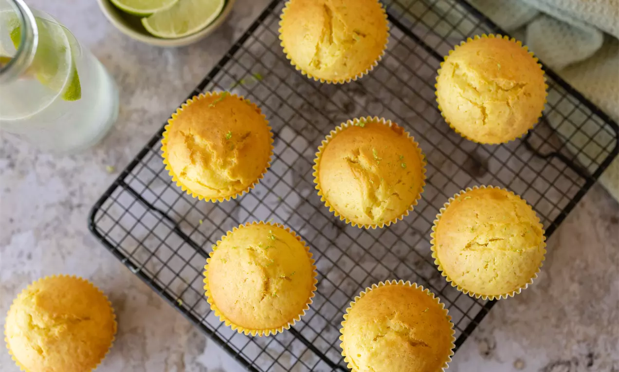 Muffins dorés au citron avec des sommets bombés, disposés sur une grille noire, accompagnés de rondelles de citron vert et d'une carafe d'eau.