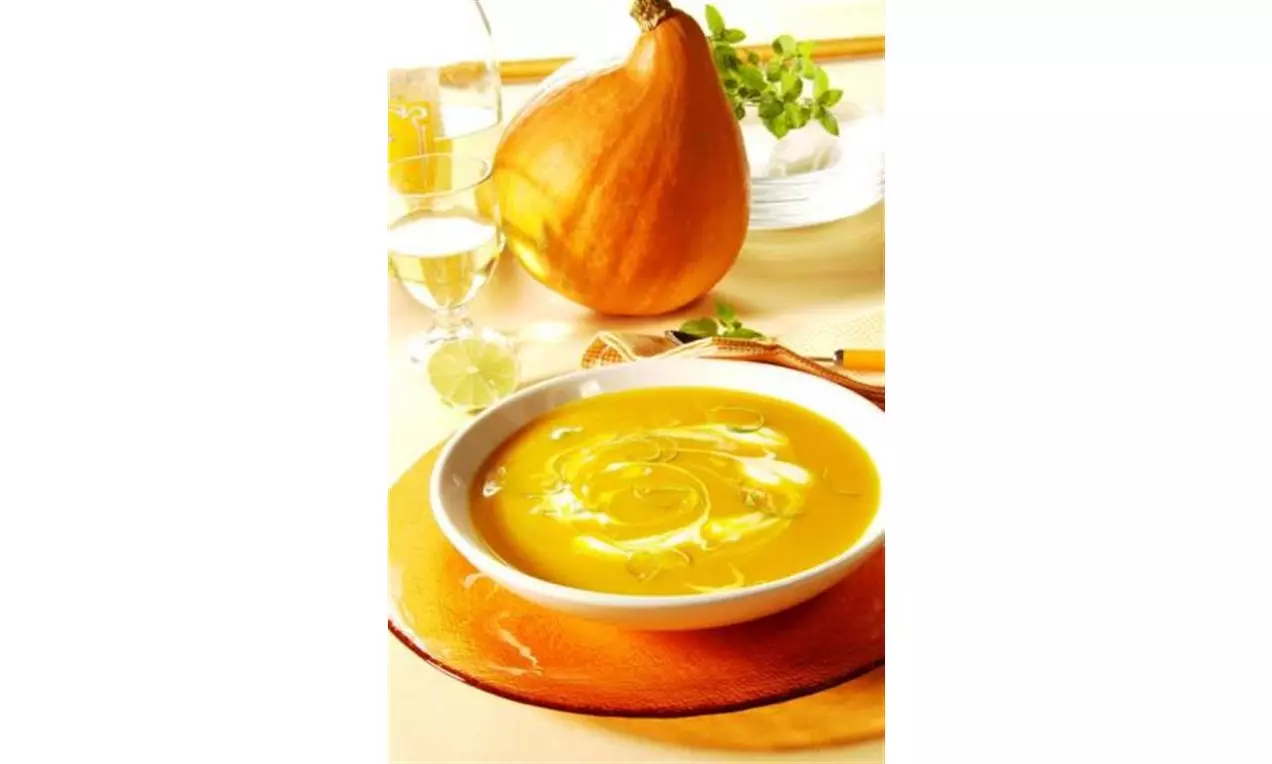 Potage onctueux orange lumineux dans une assiette blanche, décoré de fines spirales, posé sur un napperon orange avec une citrouille en arrière-plan