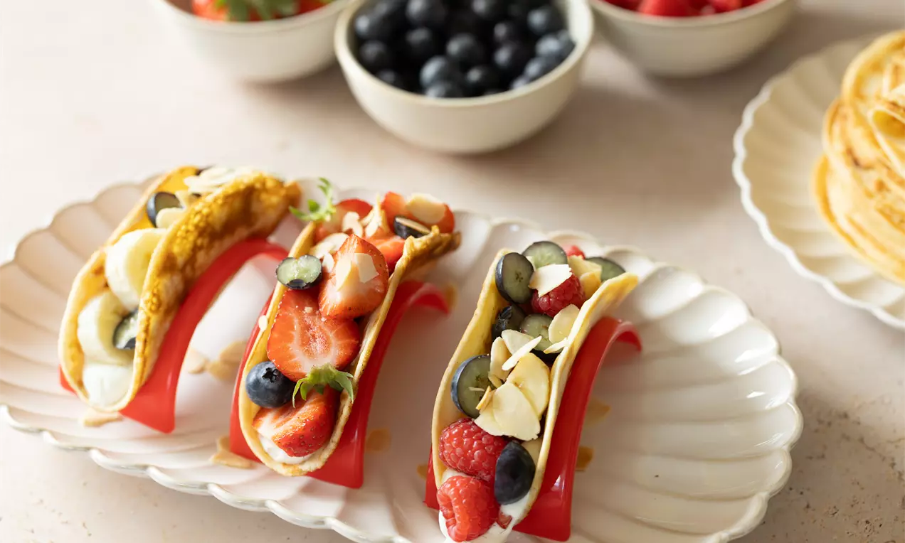 Crêpes dorées pliées en forme de tacos garnies de crème blanche, fruits rouges, myrtilles et amandes, disposées sur un plateau élégant