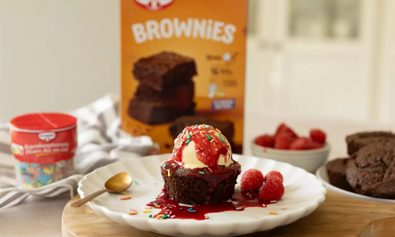 Brownie moelleux brun foncé nappé de coulis rouge, boule de glace vanille et vermicelles colorés, accompagné de framboises sur assiette blanche