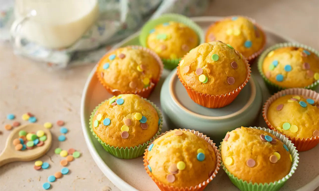 Goudgeel gebakken muffins in felgekleurde bakvormpjes, bestrooid met kleurrijke spikkels, gepresenteerd op een wit bord met decoratieve beker