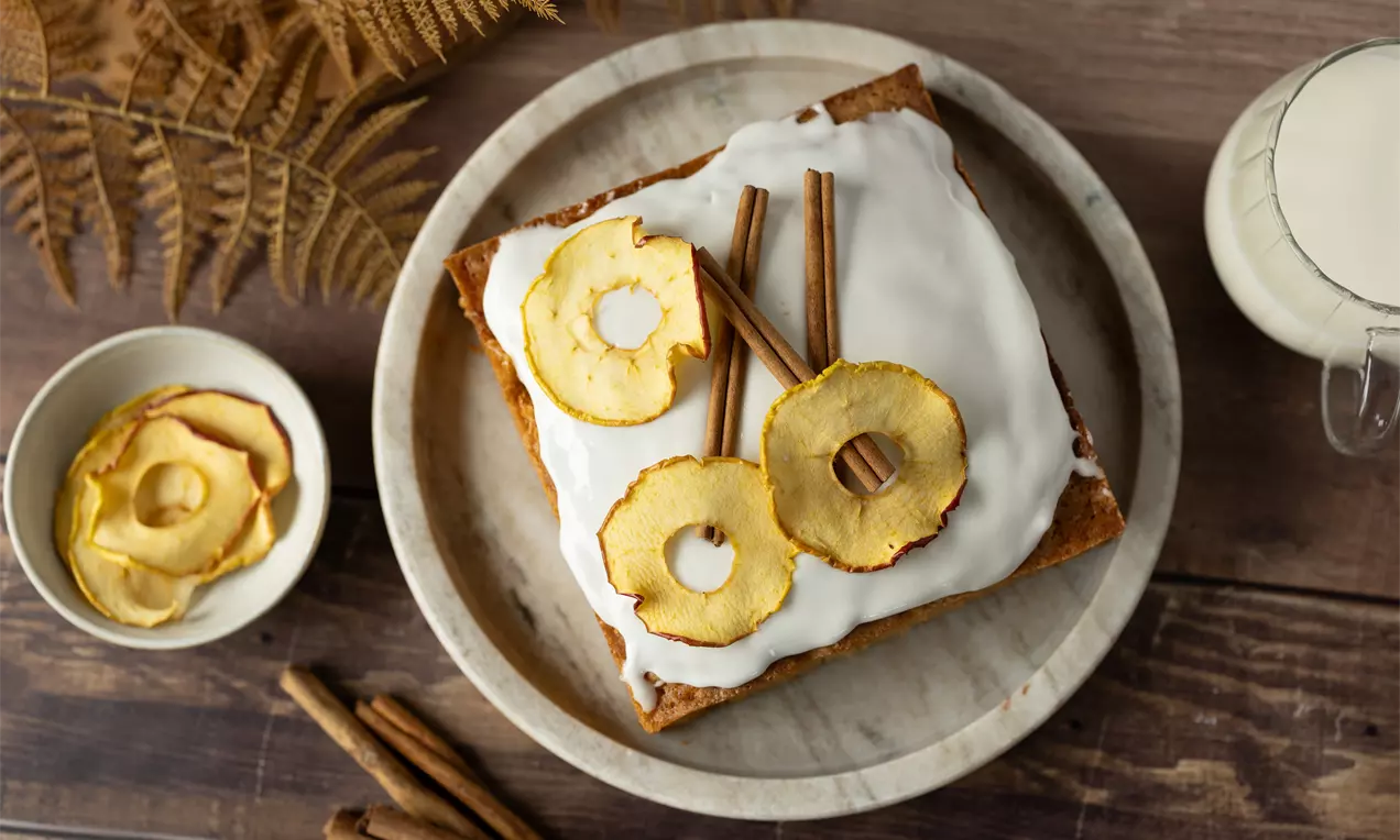 Vierkante cake met witte glazuurlaag, gegarneerd met gedroogde appelringen en kaneelstokjes op een ronde stenen schaal