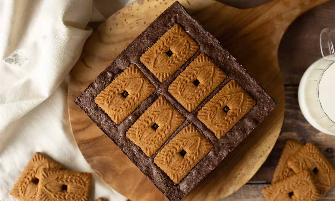 Carré de brownie chocolaté foncé avec six biscuits dorés Lotus disposés en grille sur une planche ronde en bois.