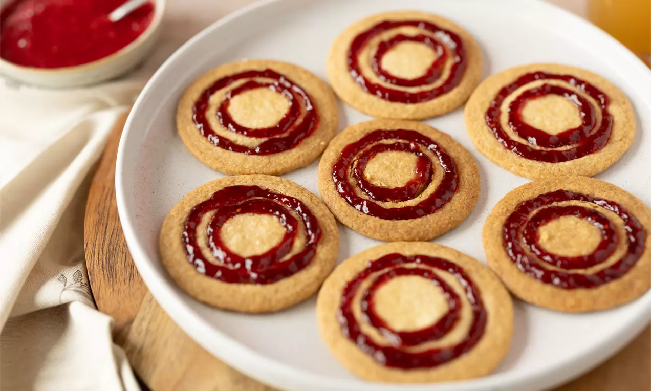 Goudbruine koekjes met een rode jamcirkel en een lichtgekleurde kern op een wit bord, geserveerd op een houten ondergrond met een jampotje