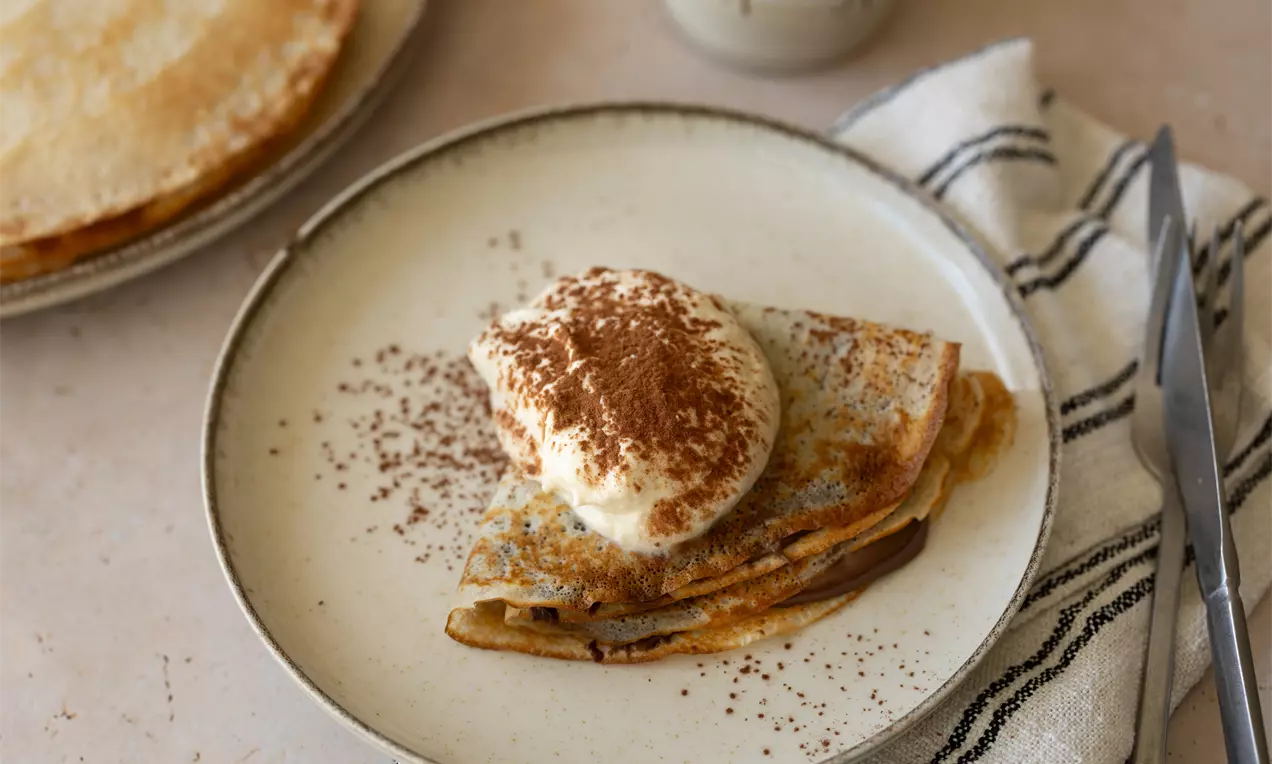 Een opgevouwen pannenkoek met lichtbruine korst, besprenkeld met cacaopoeder en een toef slagroom, geserveerd op een wit bord met bestek