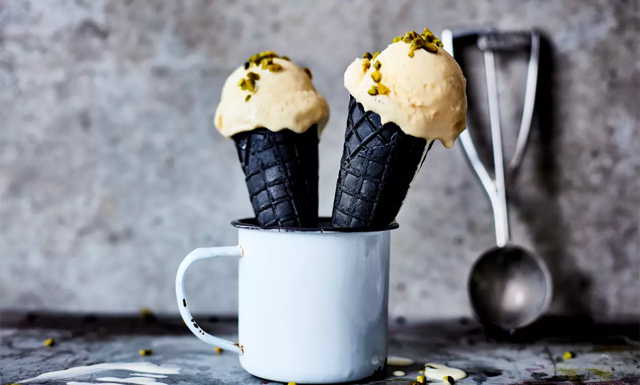 Glace à la vanille crème pâle sur des cornets noirs gaufrés, parsemée de pistaches broyées, présentée dans une tasse blanche métallique