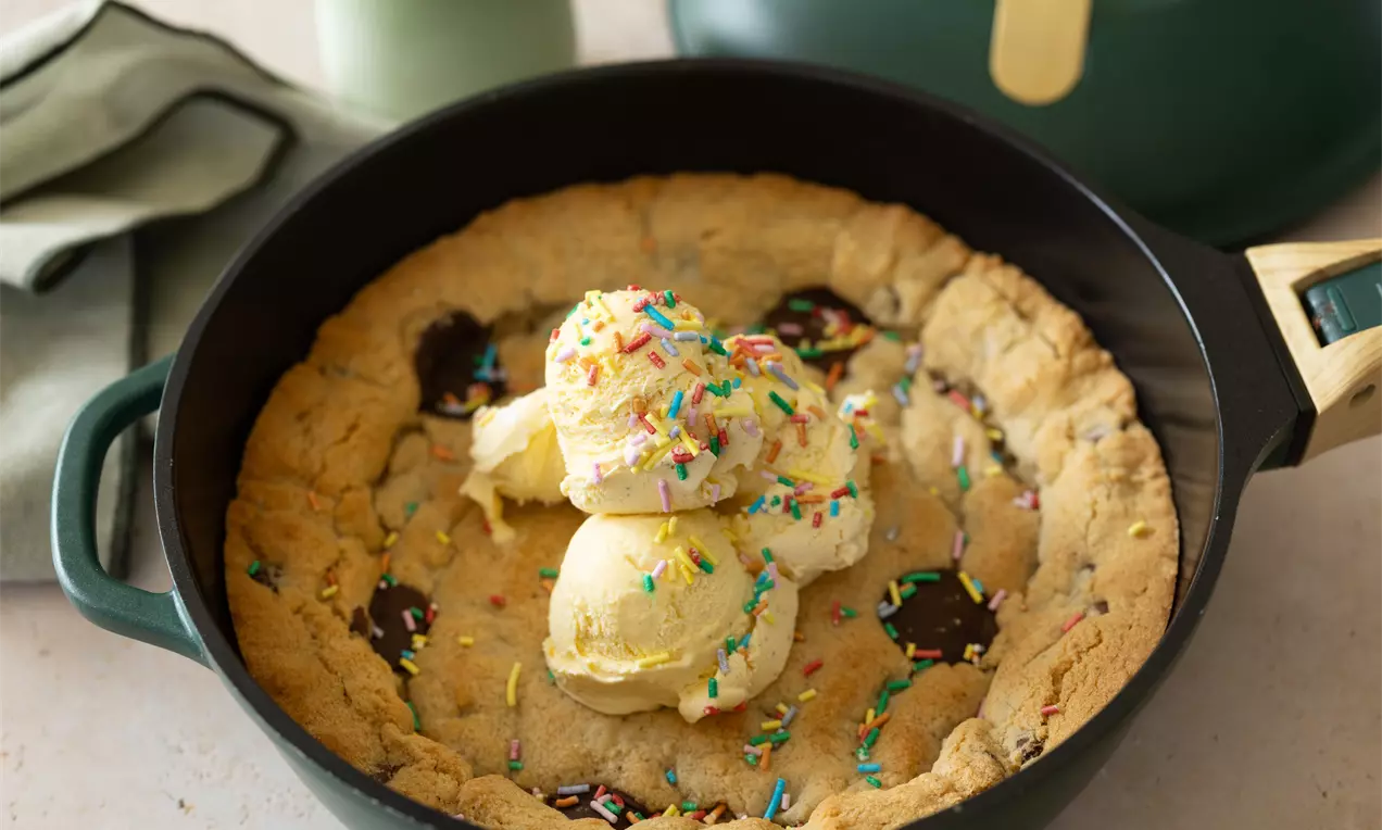 Cookie doré et moelleux dans une poêle en fonte, garni de boules de glace vanille et de vermicelles colorés.