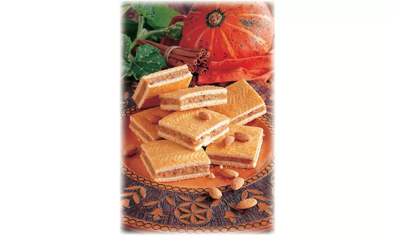 Carrés de biscuits à la citrouille dorés avec une texture ondulée, garnis d’amandes entières sur un plat décoré aux motifs chaleureux