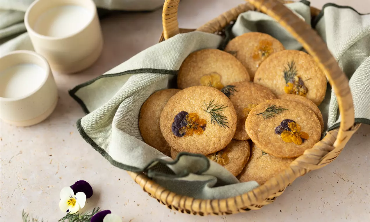 Biscuits dorés ronds décorés de fleurs comestibles et d’aneth, disposés dans un panier en osier avec un linge vert clair
