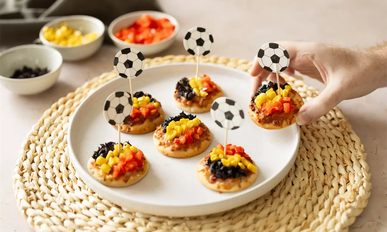 Mini-pizza’s met kleurrijke groenten, rode en gele paprika en zwarte olijven op een wit bord met voetbalprikkers, gepresenteerd op een mandvloer