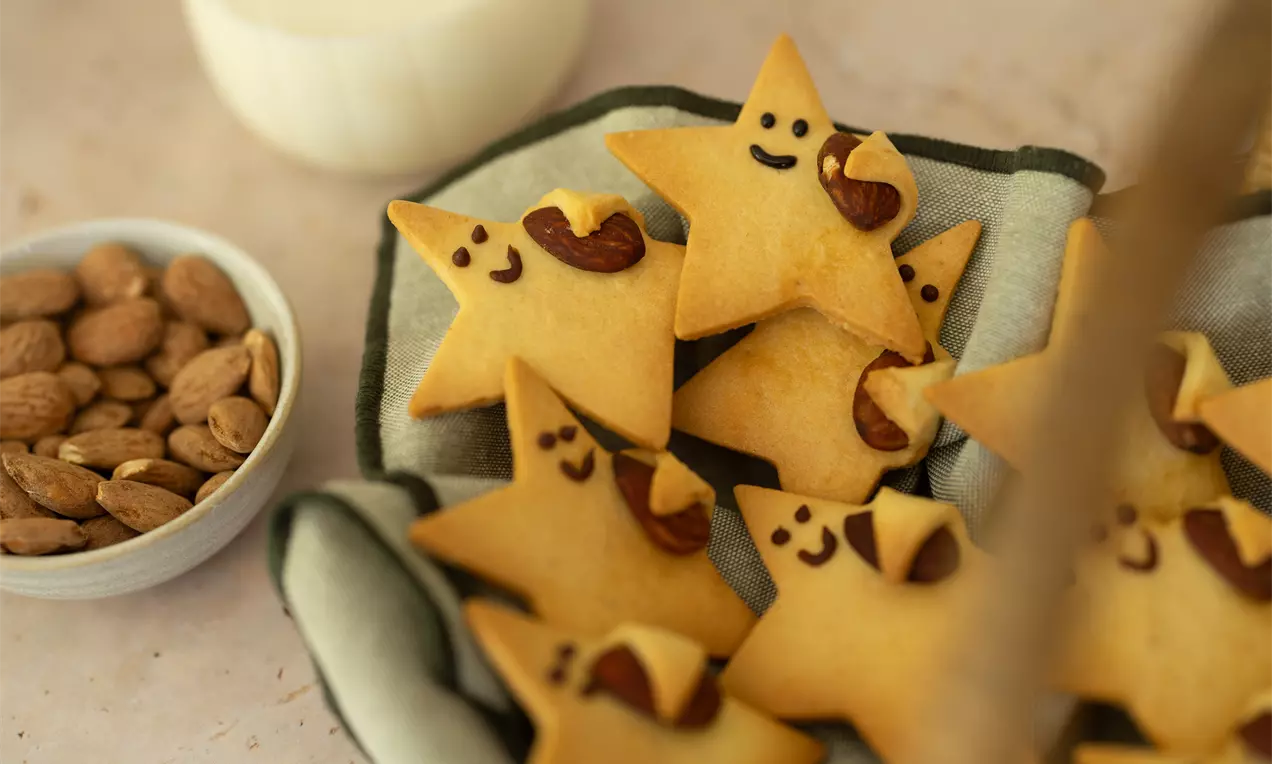 Stervormige goudbruine koekjes met getekende gezichtjes, amandelen en een vleugje chocolade, geserveerd op een licht doek met een schaaltje noten