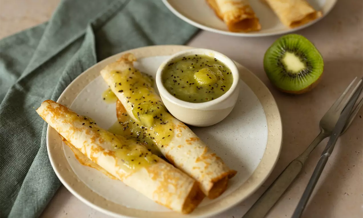 Goudbruine opgerolde pannenkoeken met glanzende kiwi-saus, geserveerd op een beige bord met een schaaltje saus, kiwi en bestek