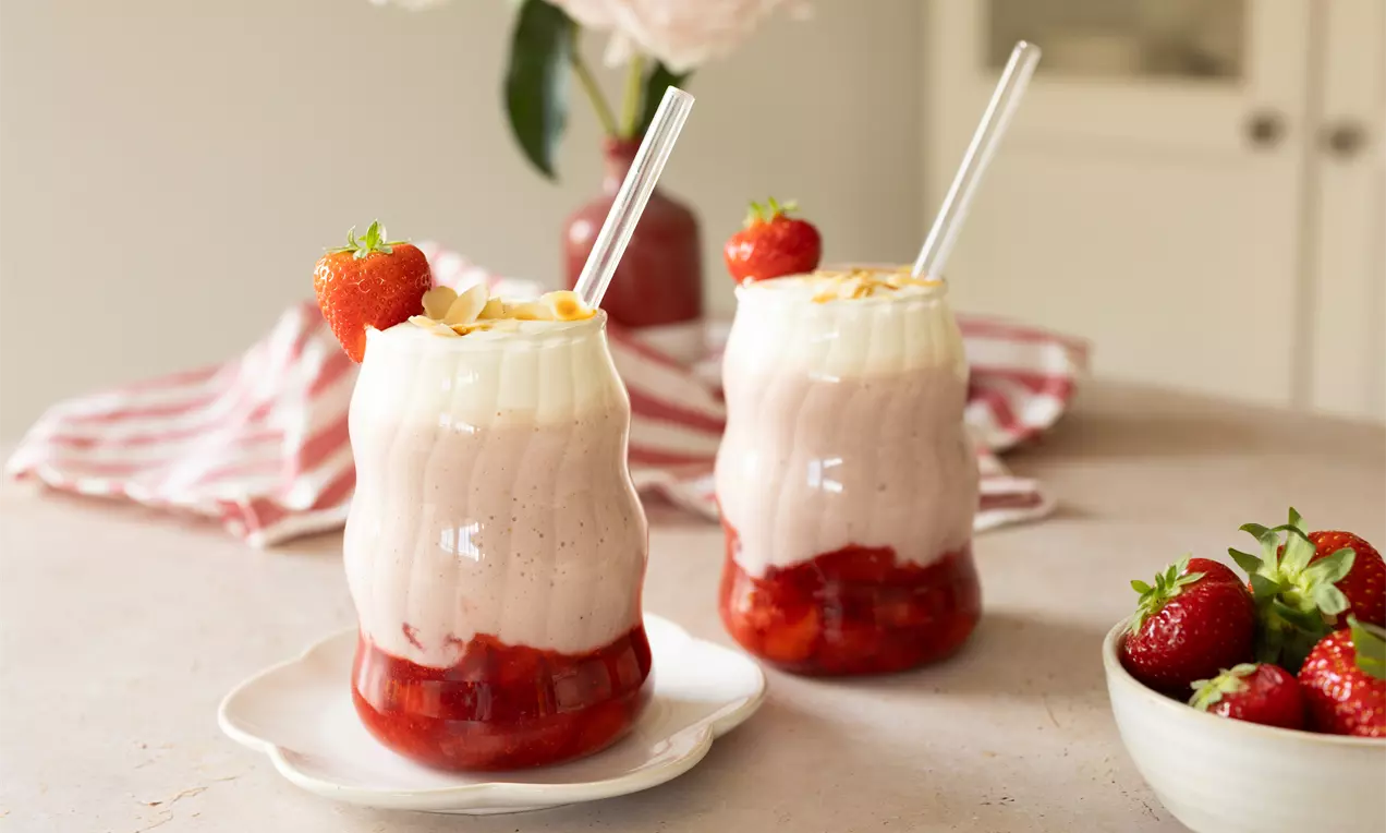 Twee gegolfde glazen gevuld met roze aardbeienmilkshake, rode coulis, slagroom en aardbeien, geserveerd op een bord naast verse aardbeien