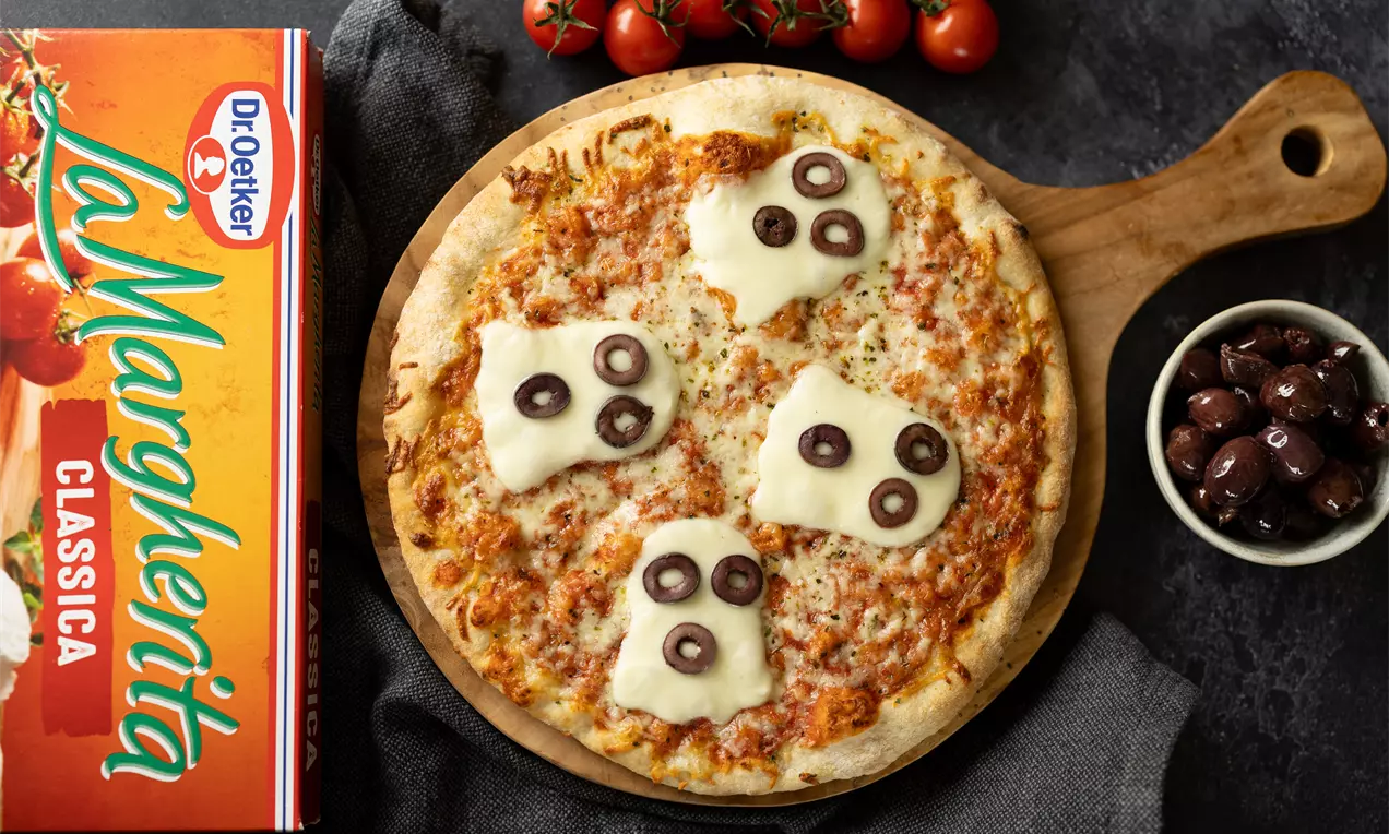 Margherita-pizza met gesmolten kaas in spookvorm en plakjes zwarte olijf op een houten serveerplank, naast tomaatjes en olijven
