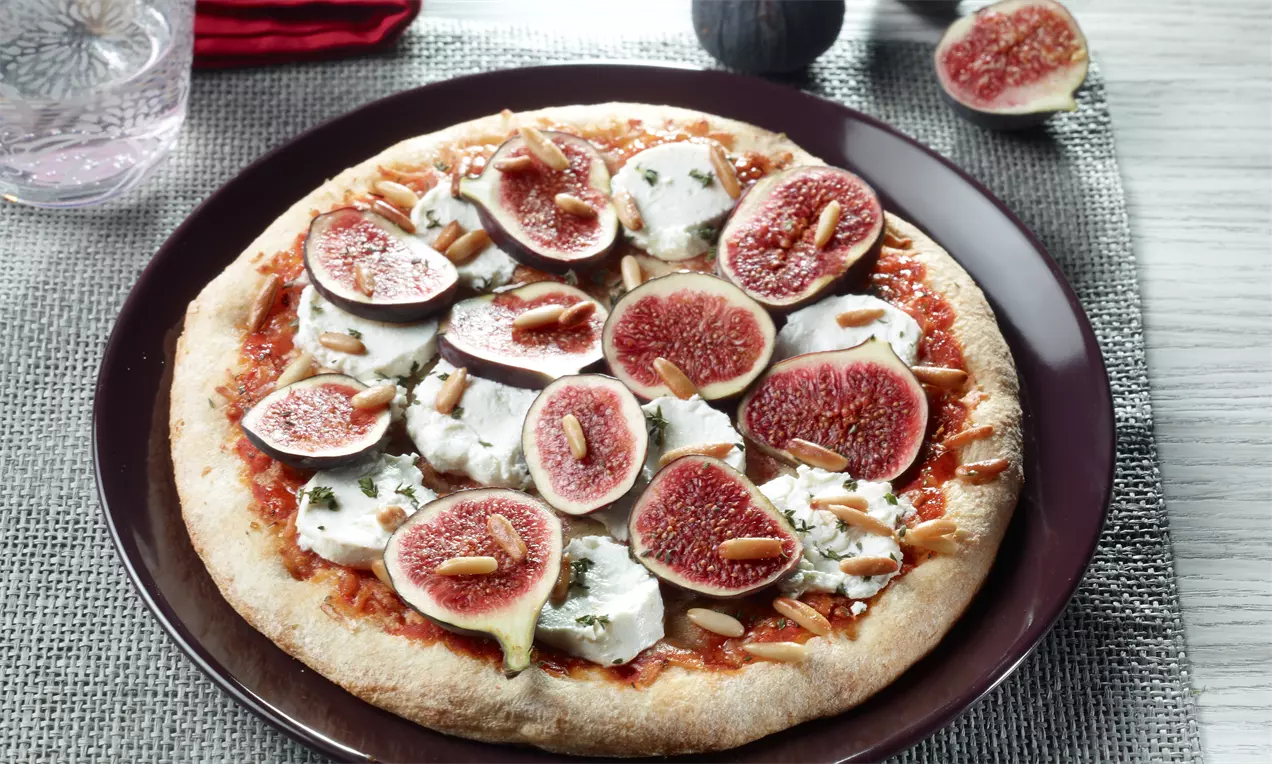 Ronde pizza op een donker bord met vijgenhelften, romige geitenkaas, pijnboompitten en rode tomatensaus op een goudbruine korst