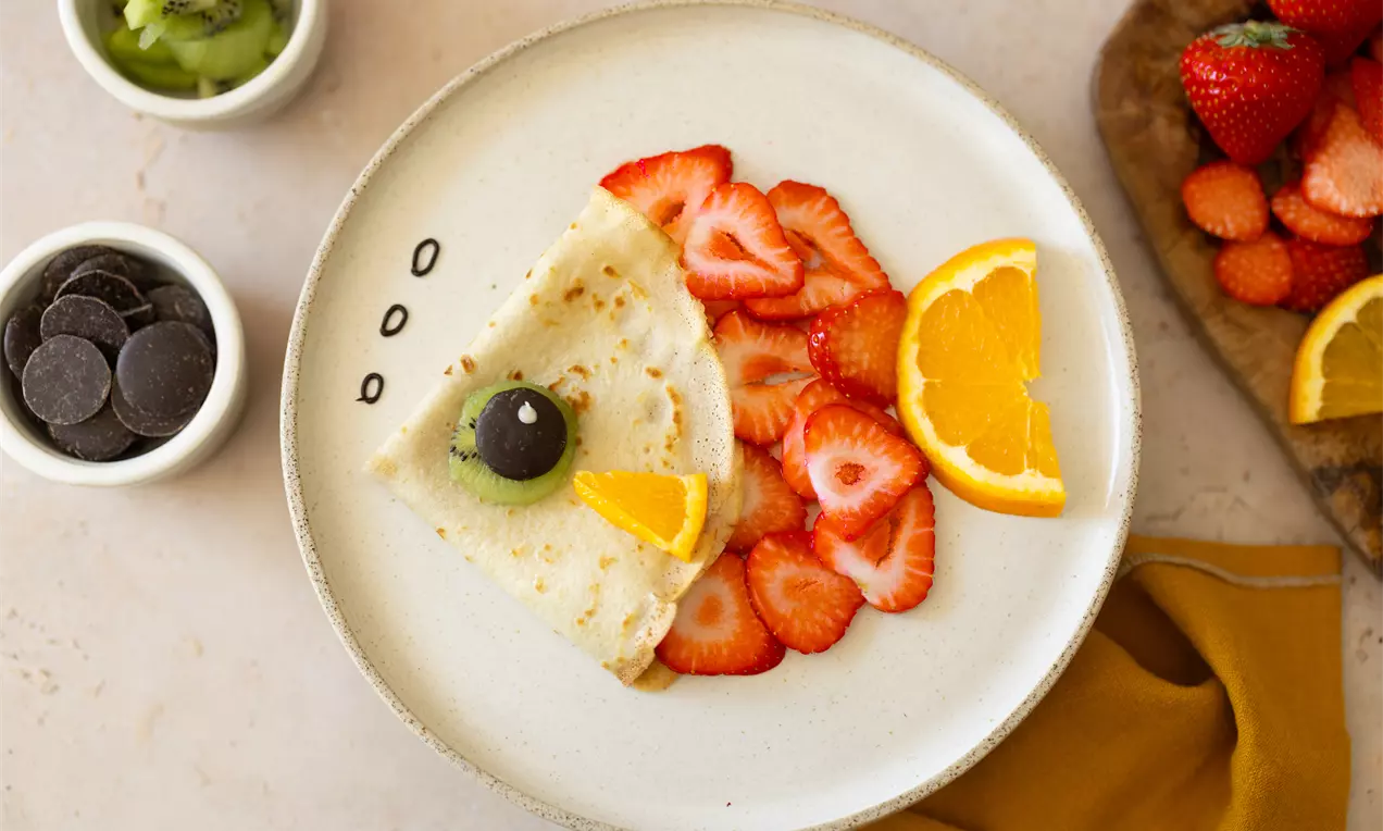 Crêpe pliée garnie d’un kiwi et d’un raisin noir, entourée de tranches de fraises et d’une rondelle d’orange sur une assiette blanche