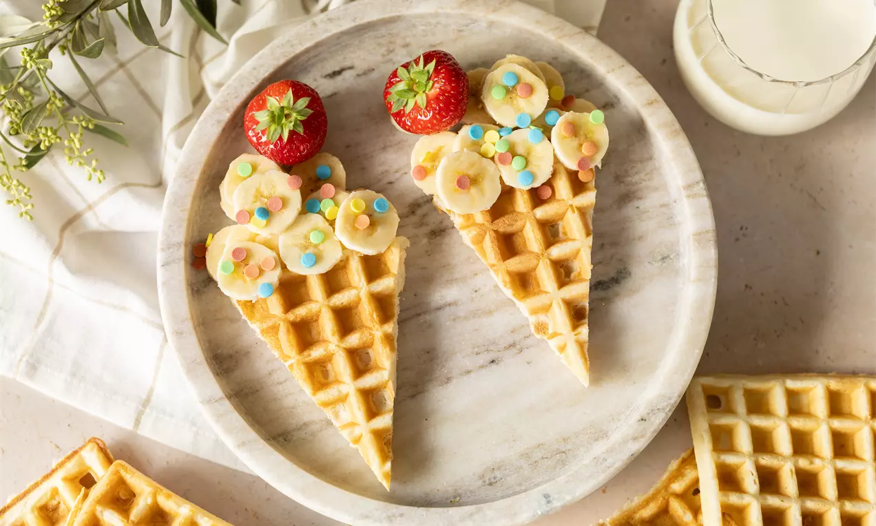 Gaufres en triangle garnies de rondelles de banane, fraises rouges et perles colorées sur une assiette marbrée avec du lait à côté