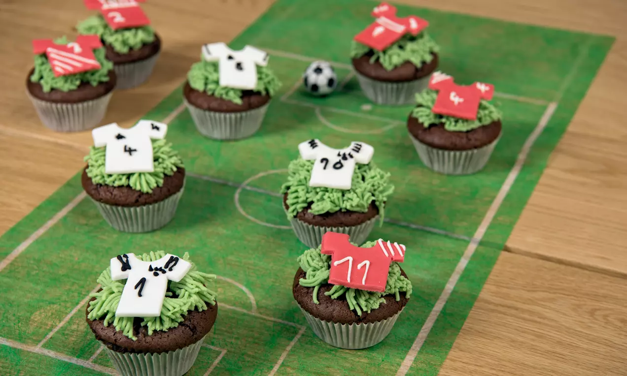 Chocolademuffins met groene crèmegrasdecoratie en fondantvoetbalshirts in rood en wit, geplaatst op een speelveldservet