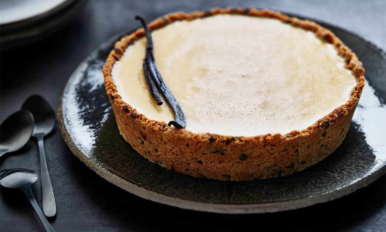Tarte ronde à la croûte dorée et granuleuse, garnie d’une crème lisse vanillée beige clair, décorée de gousses de vanille noires
