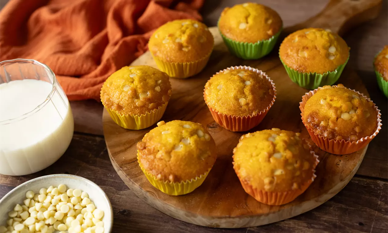Goudgele muffins in kleurrijke papiertjes met een lichte topping van witte chocoladestukjes op een houten plank, omringd door melk en een schaaltje chocola