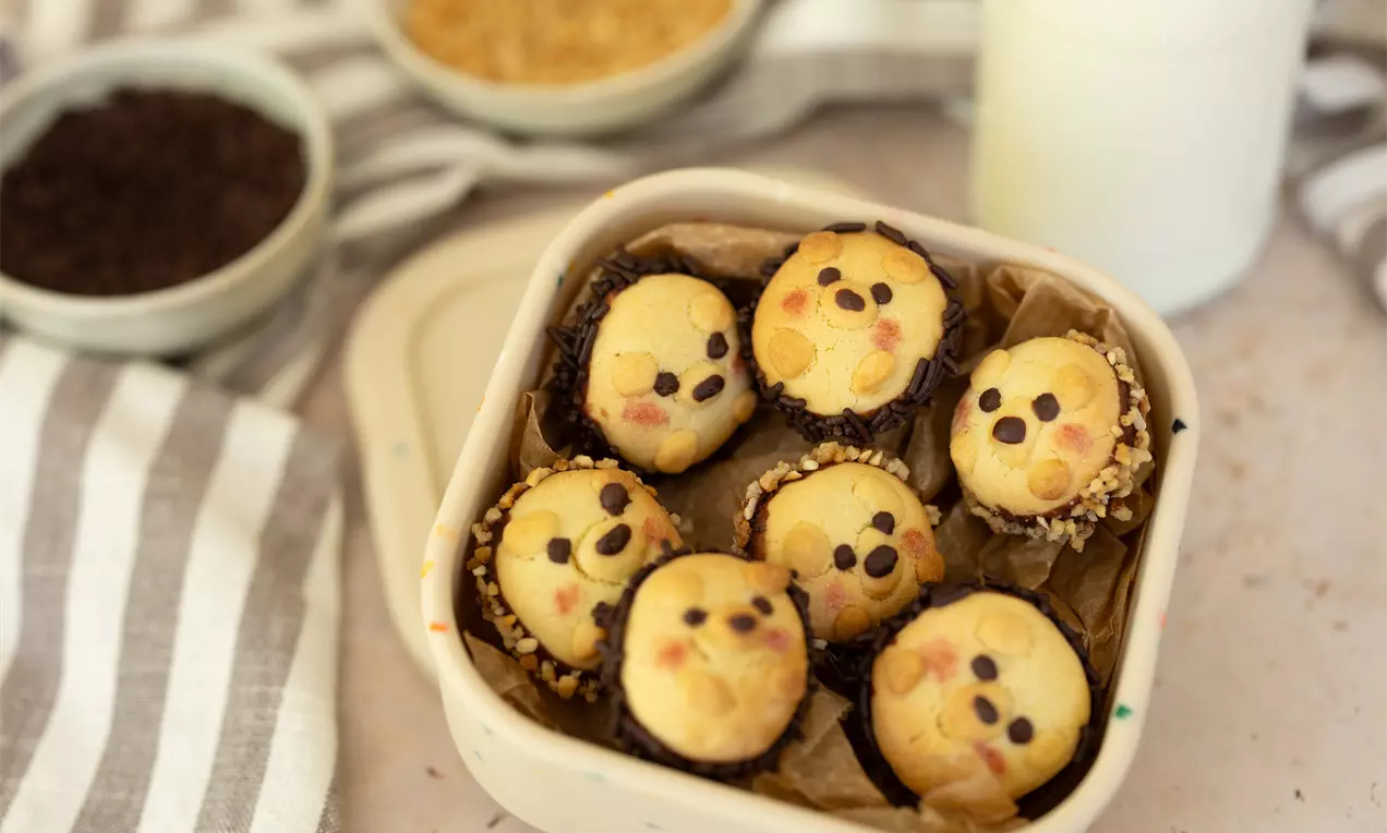 Lichte bolvormige koekjes met chocoladestukjes en getinte glazuurgezichtjes, gerangschikt in een bakje met bruine randen van papier