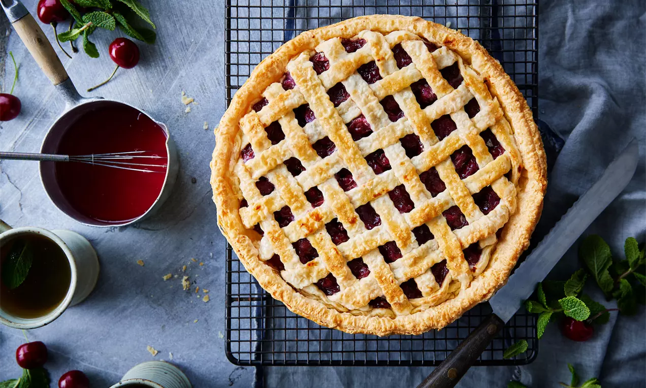 Tarte dorée avec une pâte en croisillons laissant entrevoir des cerises juteuses rouges, posée sur une grille avec des feuilles de menthe et des ustensiles