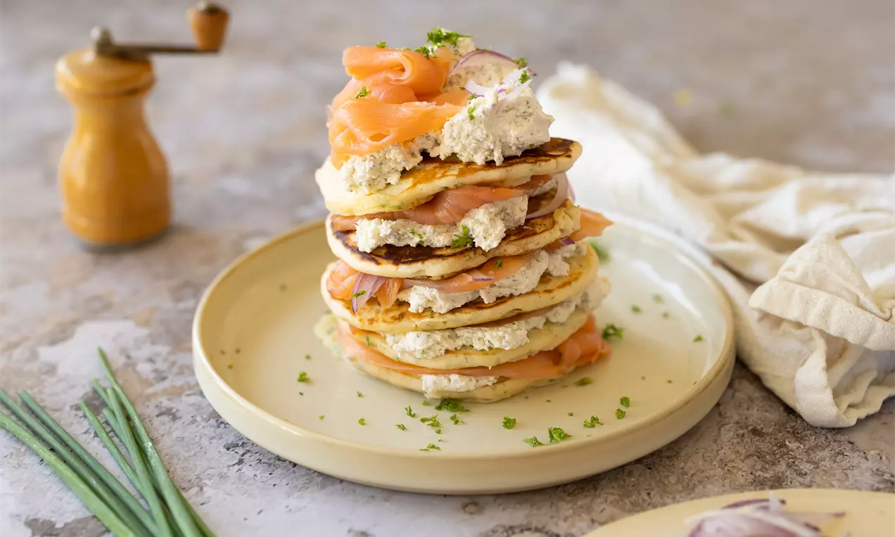 Pancakes dorés empilés garnis de tranches de saumon rose, crème blanche et oignons rouges, parsemés de ciboulette verte sur une assiette claire