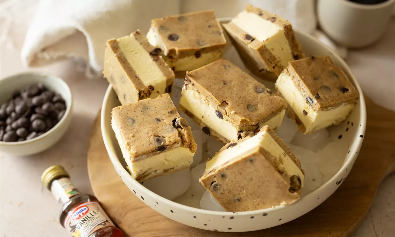 Vierkante ijscreaties tussen lichtbruine koekjes met chocoladestukjes op een wit stippenbord, omringd door vanille-extract en chocoladedecoratie