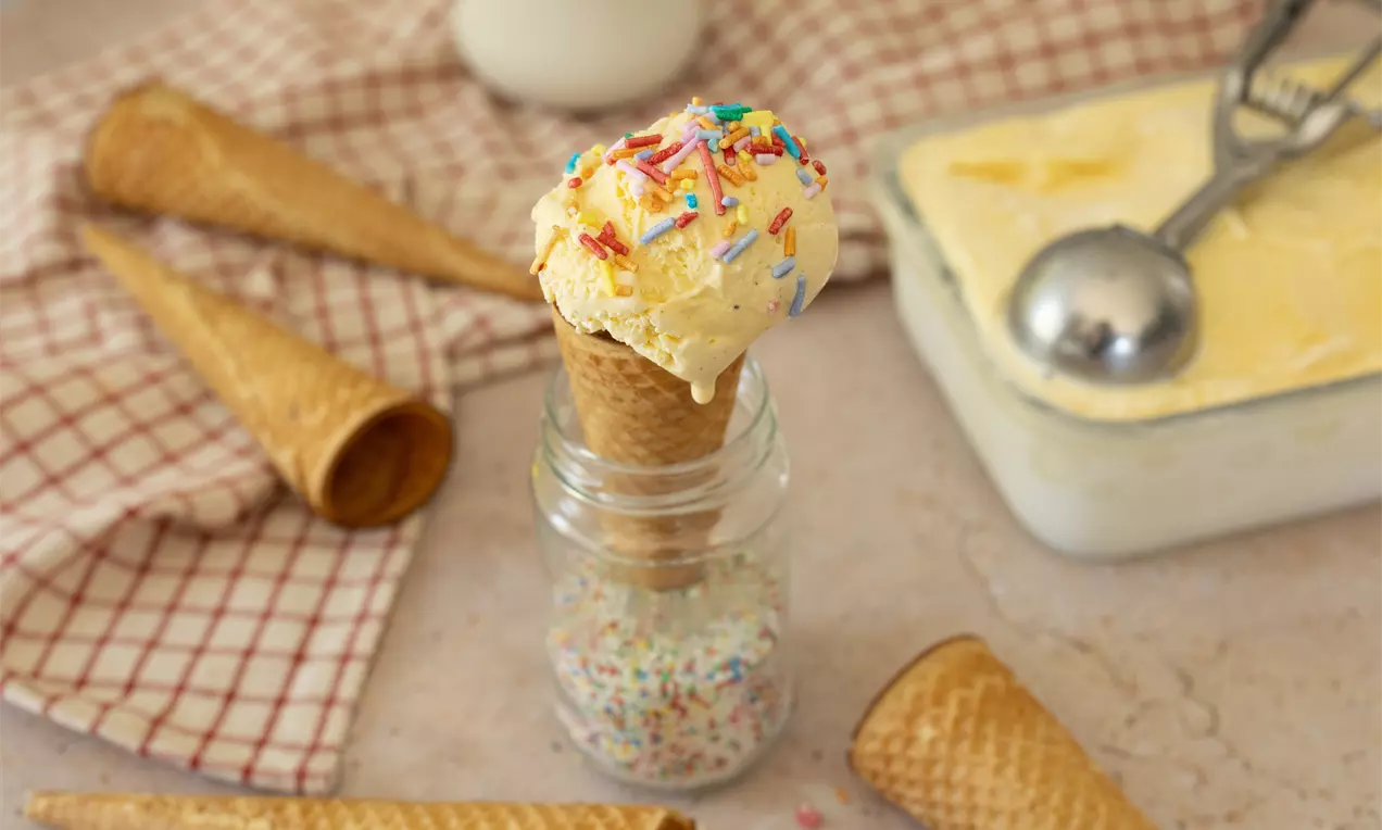 Vanille-ijs met kleurrijke sprinkels in een hoorntje, staand in een glazen pot met sprinkels, omgeven door hoorntjes en ijsbak op een keukenhanddoek