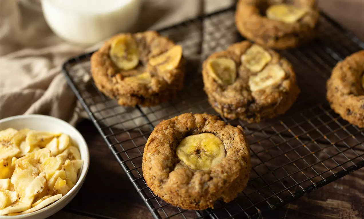 Pâtisseries dorées moelleuses avec une tranche de banane au centre, disposées sur une grille noire avec des chips de banane en accompagnement