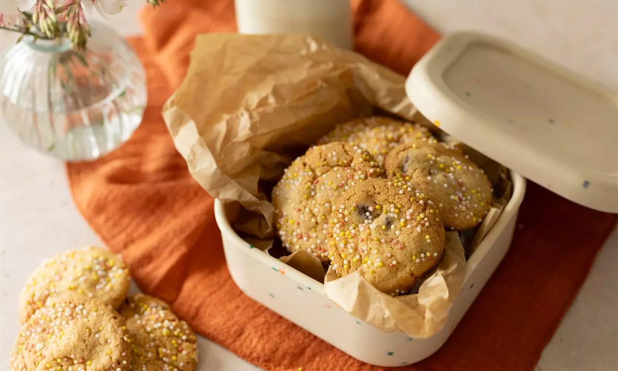 Goudbruine koekjes met sprinkles in een bakje met bakpapier, op een oranje doek met een melkglas en bloemenvaasje op de achtergrond
