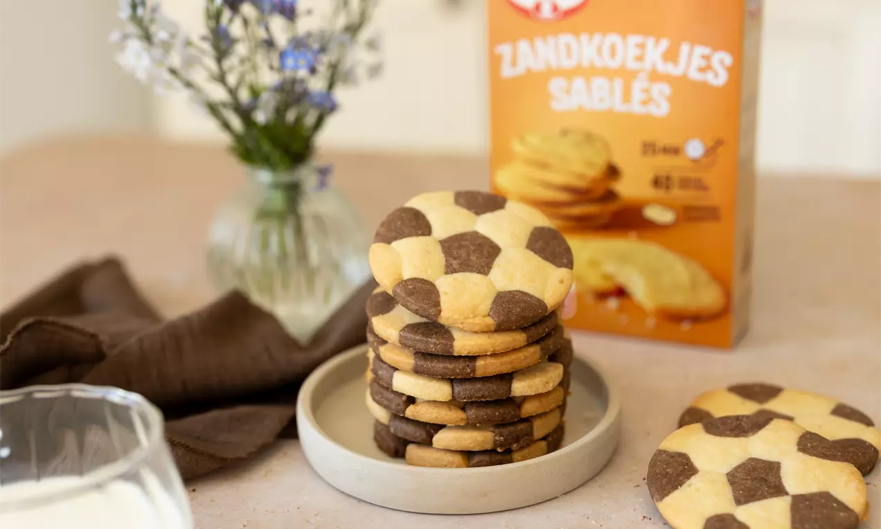 Biscuits sablés ronds jaune clair et marron disposés en pile sur une assiette beige, avec un verre de lait et une fleur en arrière-plan