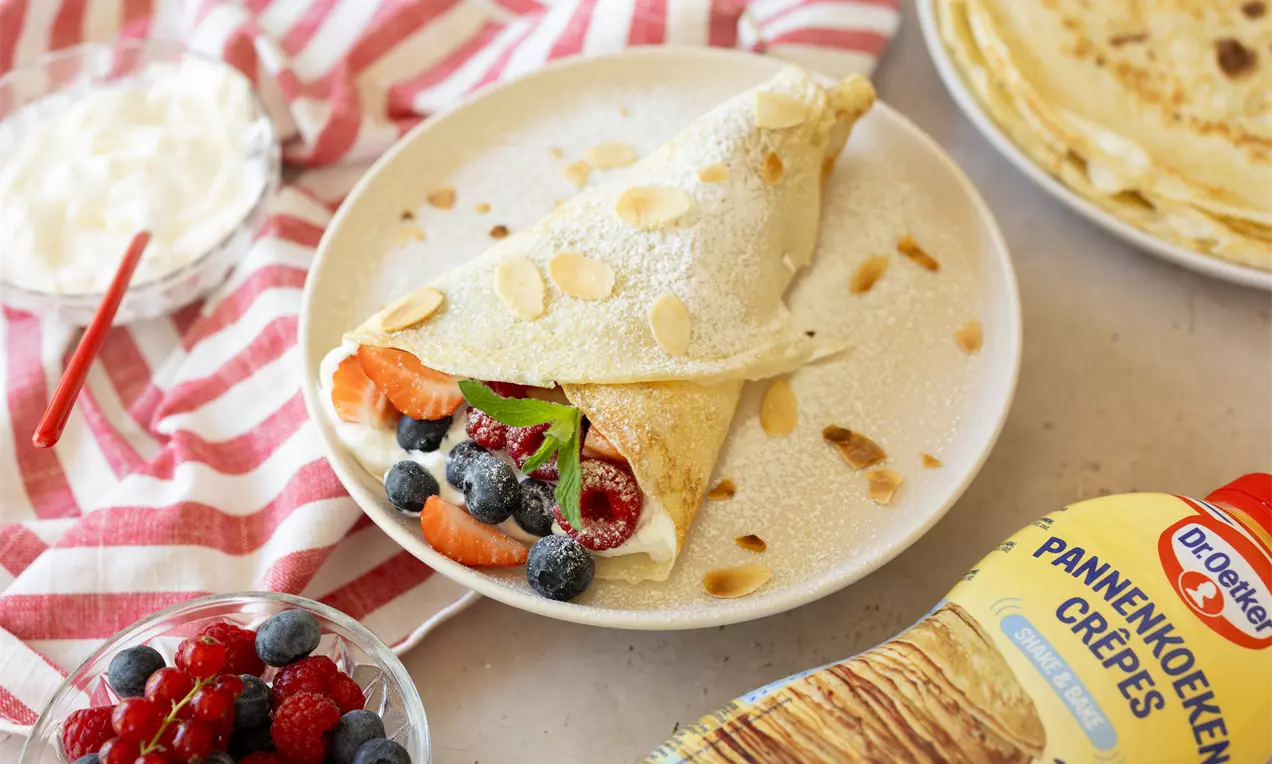 Dunne goudgele pannenkoek met frisse bessen en room in een hoorntje, bestrooid met amandelschaafsel op een wit bord met rood gestreepte doek