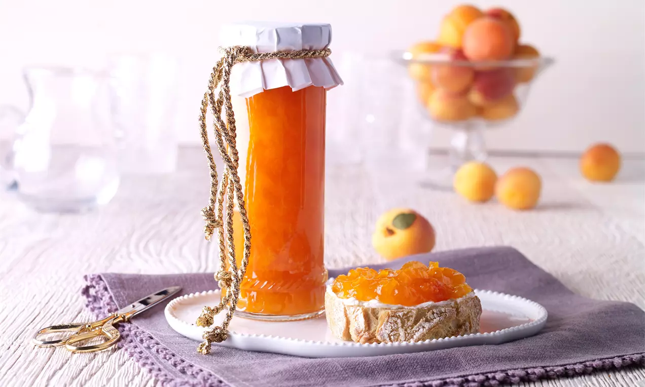 Confiture d'abricots dorée et brillante dans un pot décoré, posée sur une assiette blanche avec pain couvert de confiture sur une nappe lavande