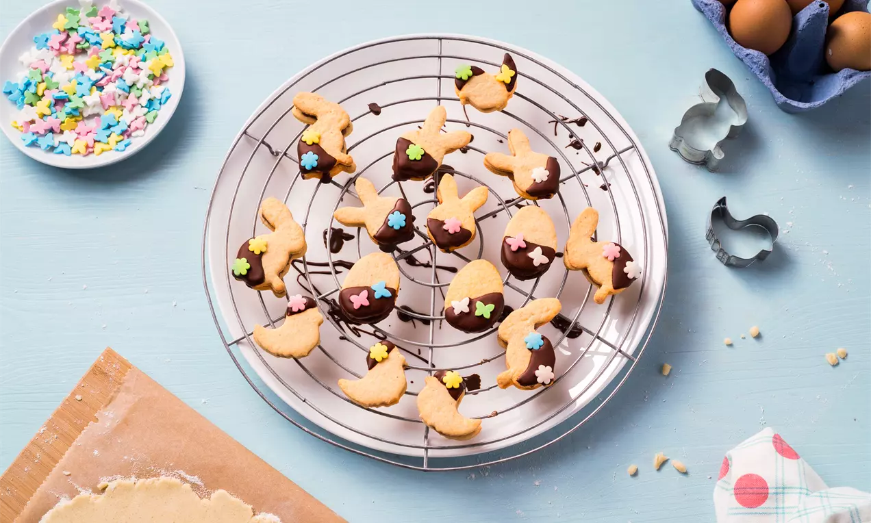 Biscuits de Pâques dorés en formes de lapins et poussins nappés de chocolat noir, décorés de petits éclats colorés sur une grille ronde argentée