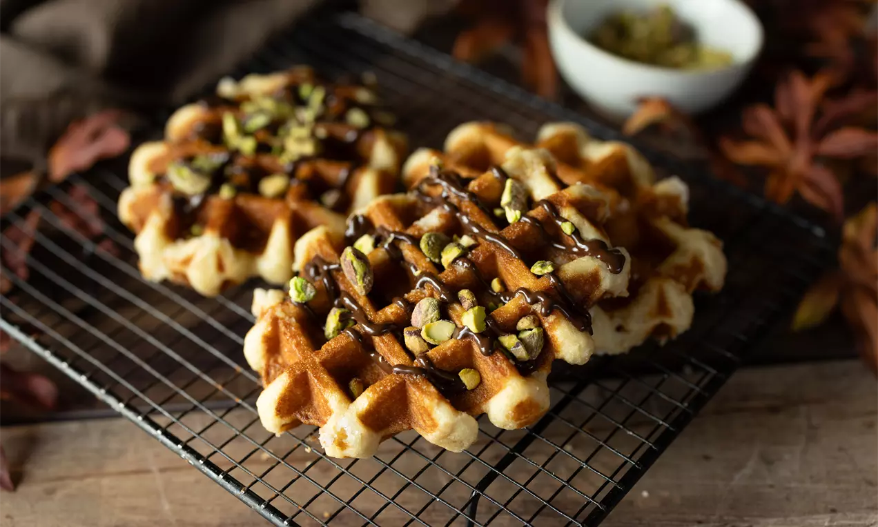Goudbruine wafels met een ruitpatroon, rijkelijk gegarneerd met bruine chocoladesaus en groene gehakte pistachenoten op een zwart rek