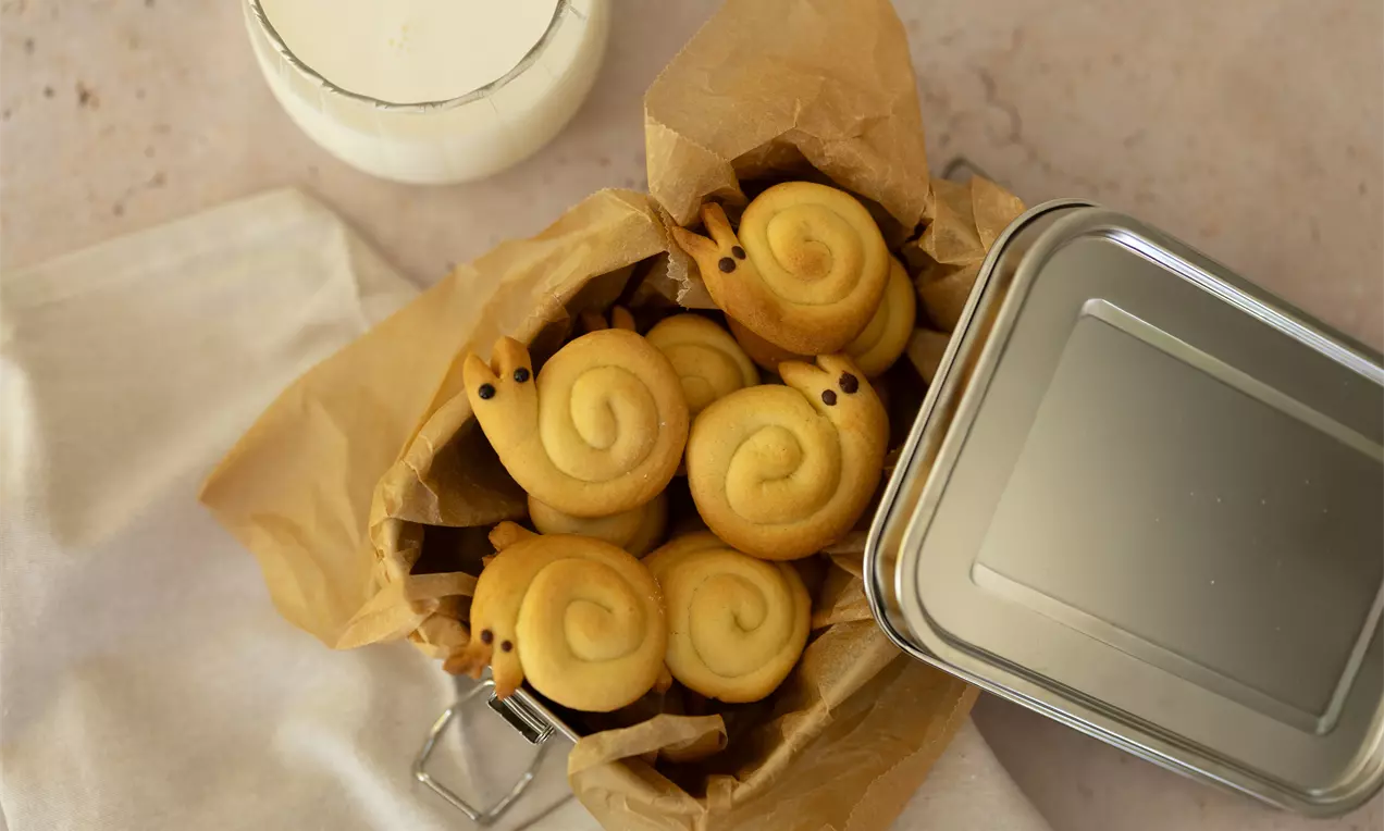 Goudbruine koekjes in de vorm van slakken met donkere oogjes, gepresenteerd in bruin bakpapier in een metalen blik naast een glas melk