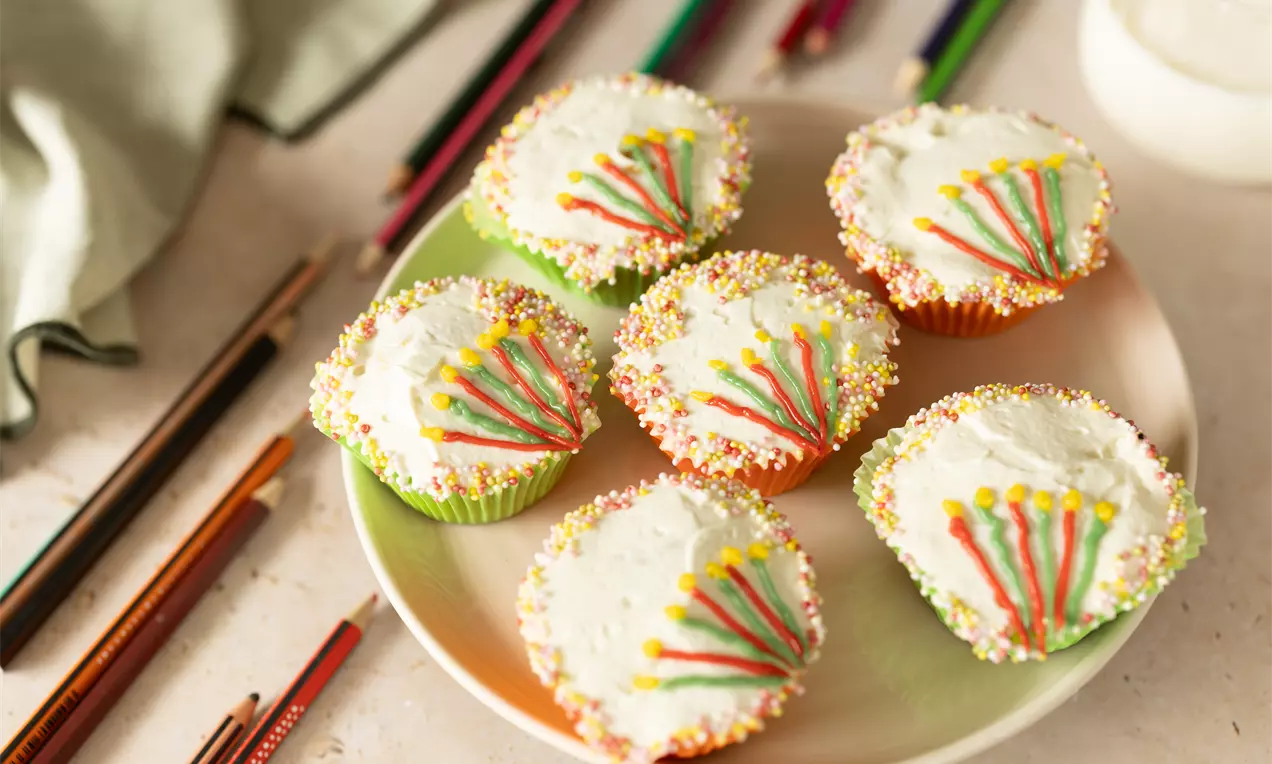 Petits cupcakes moelleux avec glaçage blanc, décorés de perles colorées et de motifs en gelée rouge et jaune sur une assiette pastel