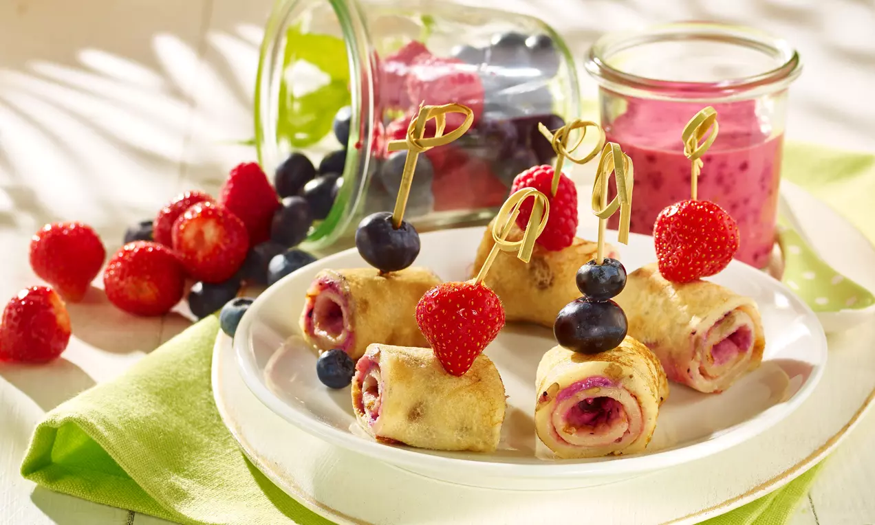Kleine opgerolde pannenkoekjes met rode vruchtenvulling, op houten prikkers met aardbeien en blauwe bessen, geserveerd op een wit bord