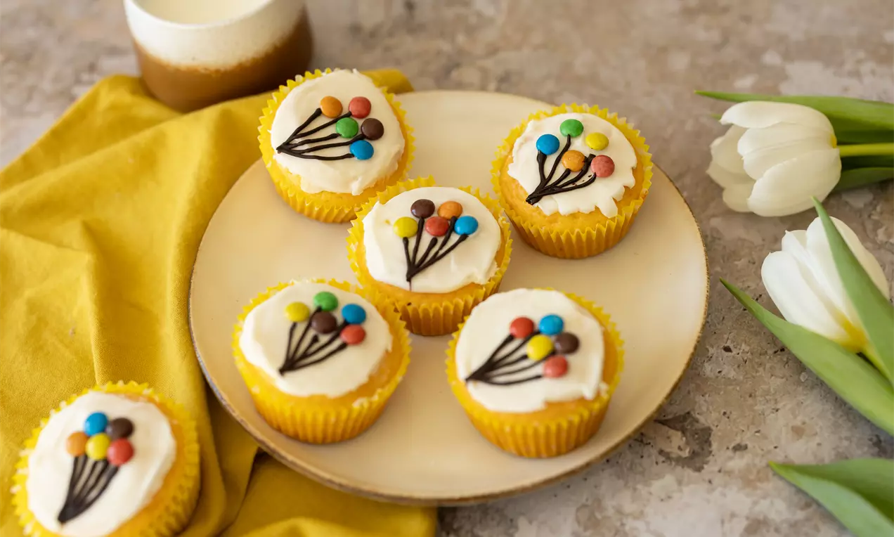 Cupcakes jaunes décorés de petits bonbons colorés en forme de ballons sur une assiette blanche, entourée de tulipes et d'un tissu jaune
