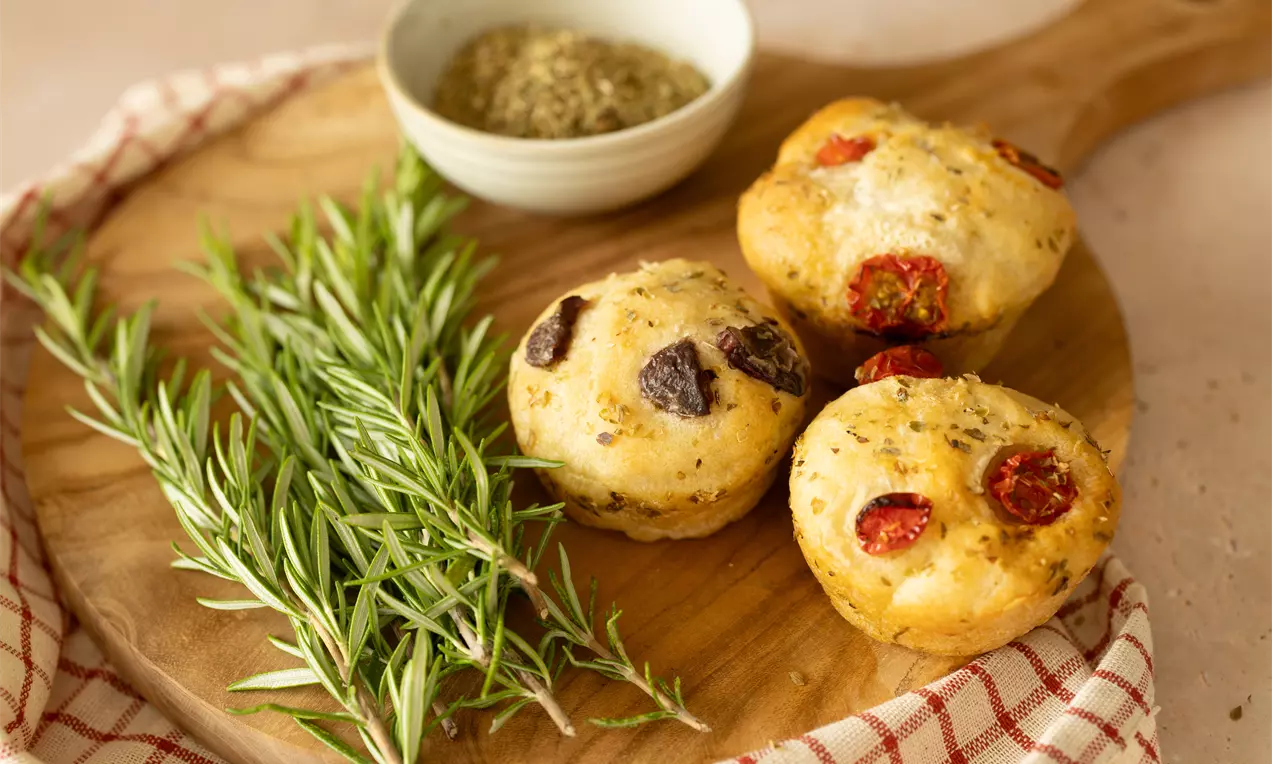 Drie goudbruine hartige muffins met tomaat en olijven op een houten plank, naast verse rozemarijntakjes en een schaaltje kruiden