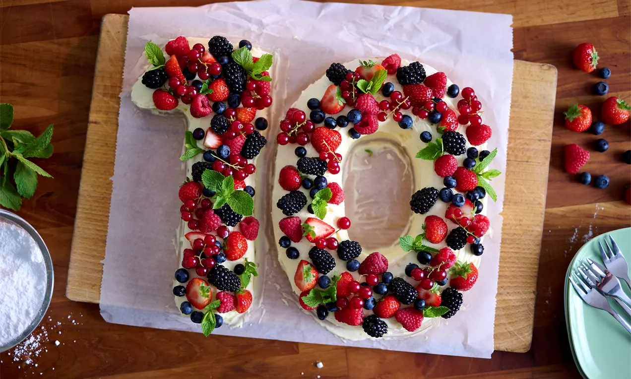 Gâteau en forme de chiffre dix, recouvert de crème légère et garni de fruits rouges brillants, de feuilles de menthe et de baies noires