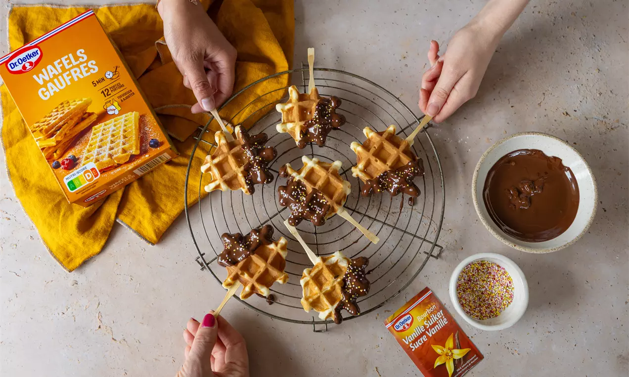 Gaufres dorées en forme de petits losanges sur bâtonnet, trempées partiellement dans du chocolat nappé de vermicelles colorés, disposées sur une grille ronde avec un bol de chocolat fondu et une touche de jaune vif apportée par un torchon plié.