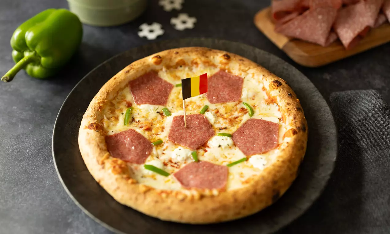 Een goudbruine pizza met salami, gesmolten kaas en dunne groene paprikareepjes, geserveerd op een zwarte schaal met Belgische vlagdecoratie